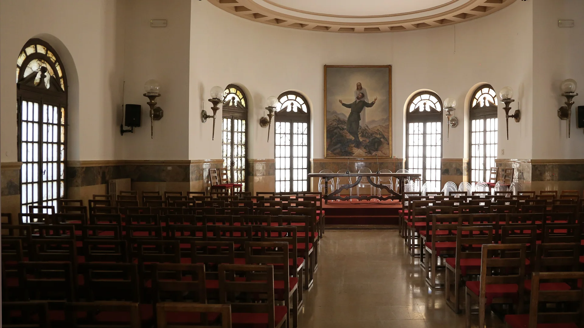 Die Sala Antonio Girelli im Palazzo dei Mutilati, Verona: Reihen von Holzstühlen mit roten Sitzen gegenüber einer geschwungenen Apsis mit Bogenfenstern und einem großen Gemälde.